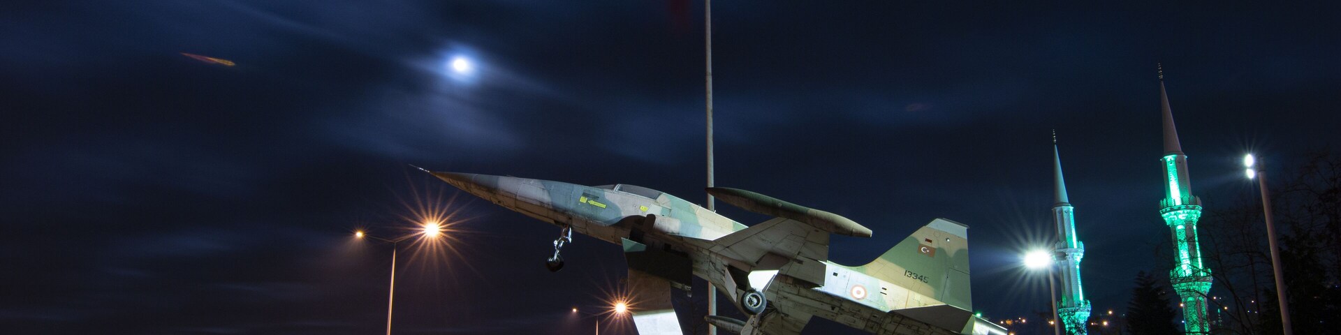 A nighttime sculpture of a military aircraft with a flagpole, illuminated by streetlights, and two green-lit towers in the background.