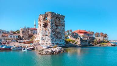 A historic stone tower by a turquoise sea, surrounded by modern buildings and boats.
