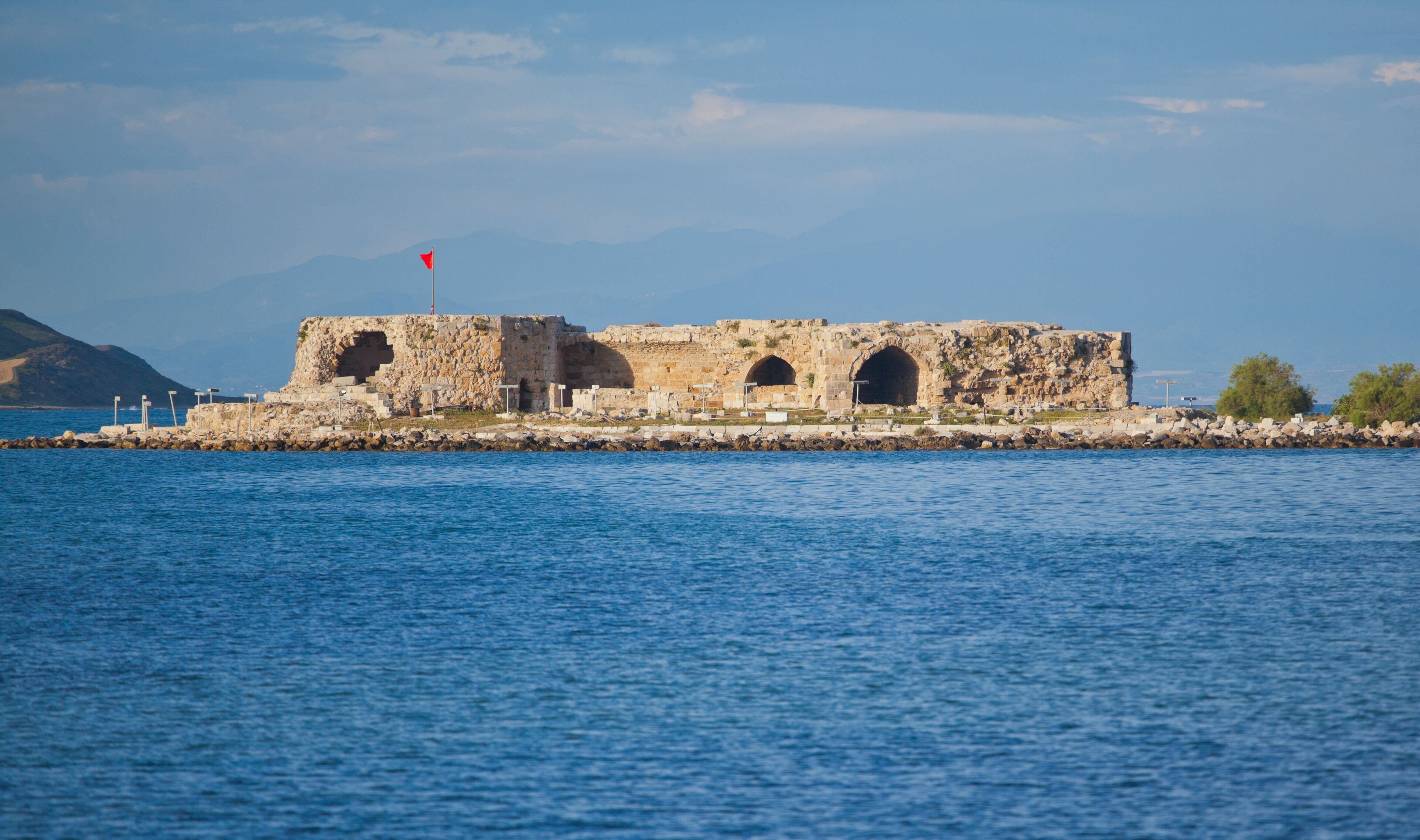 A coastal fort with three arched openings, a red flag atop, and a rocky shoreline.