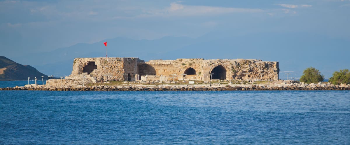 A coastal fort with three arched openings, a red flag atop, and a rocky shoreline.