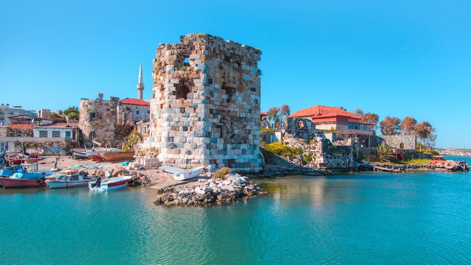 A historic stone tower by a clear blue sea, with boats docked nearby and a red-roofed building in the background.