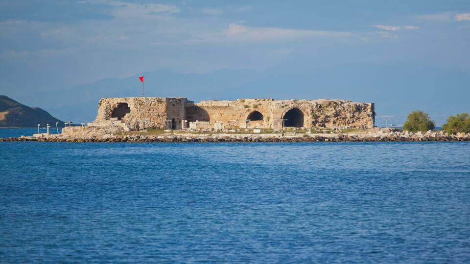 A coastal fort with three arched openings, a red flag on top, and a rocky shoreline in the background.