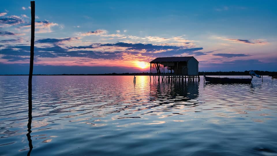 A sunset over a calm lake with a wooden dock and a small house.