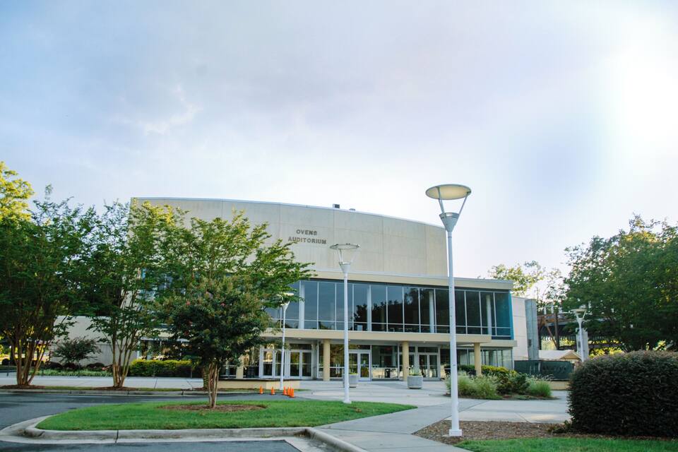 A modern building with a curved facade, labeled "OVENS AUDITORIUM," surrounded by well-maintained landscaping and a street lamp.