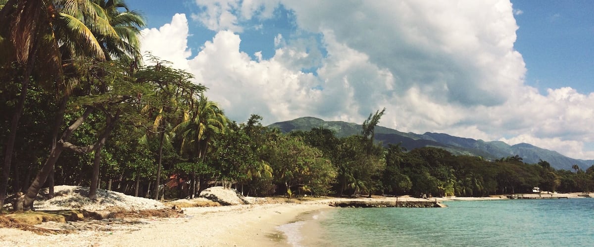 A beach with palm trees, clear turquoise water, and mountains in the background.