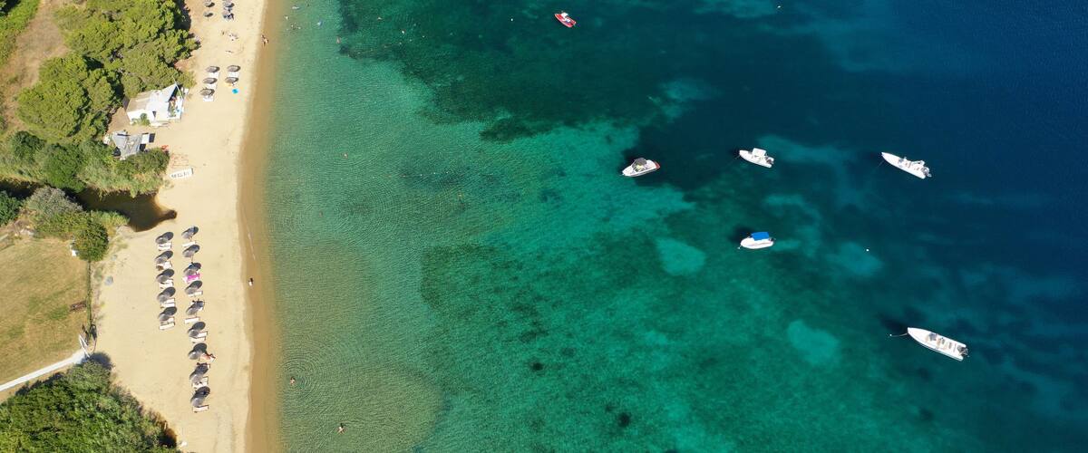 An aerial view of a beach with clear turquoise waters, several boats anchored near the shore, and a line of beach umbrellas and sun loungers along the sandy beach.