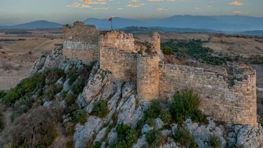 A historical stone fortress with a flag, surrounded by rocky terrain and distant mountains.