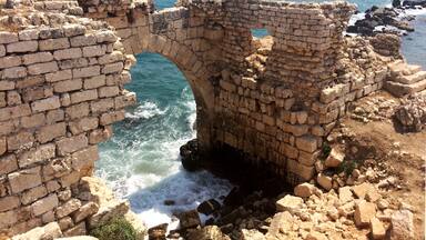 Ancient stone ruins with a large arched gateway leading to the sea, surrounded by rocky terrain and a distant fortified structure.