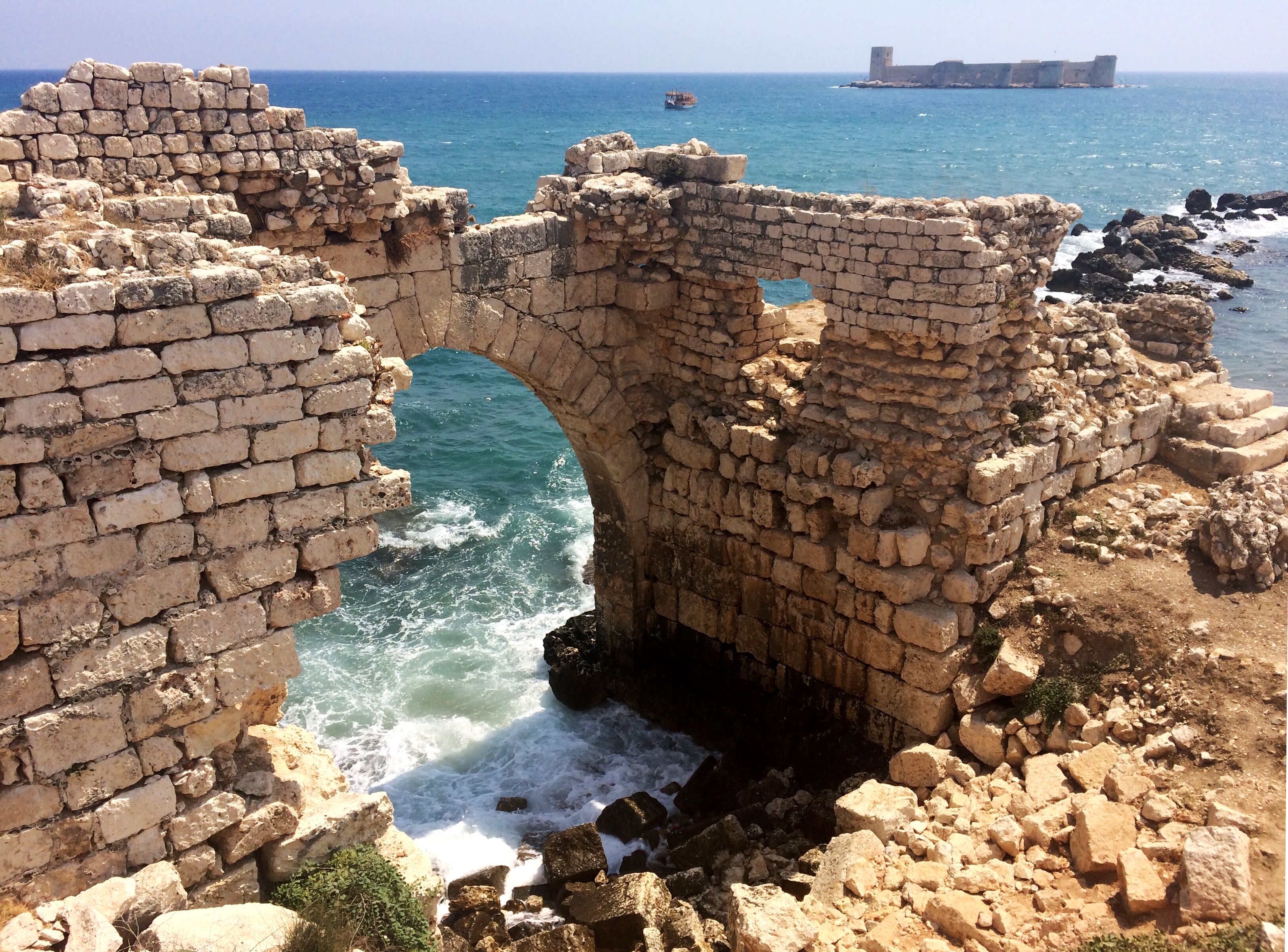 Ancient stone ruins with a large arched gateway leading to the sea, surrounded by rocky terrain and a distant fortified structure.
