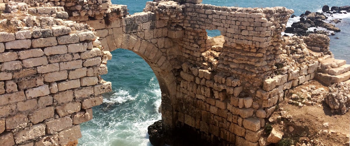 Ancient stone ruins with a large arched gateway leading to the sea, surrounded by rocky terrain and a distant fortified structure.