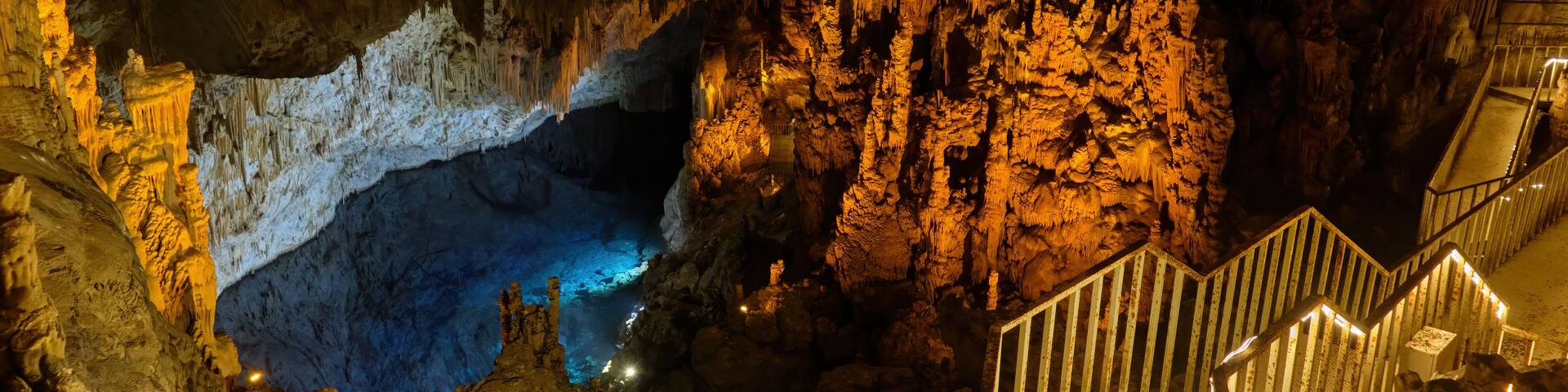 A cave with stalactites and stalagmites, a blue pool, and a staircase.