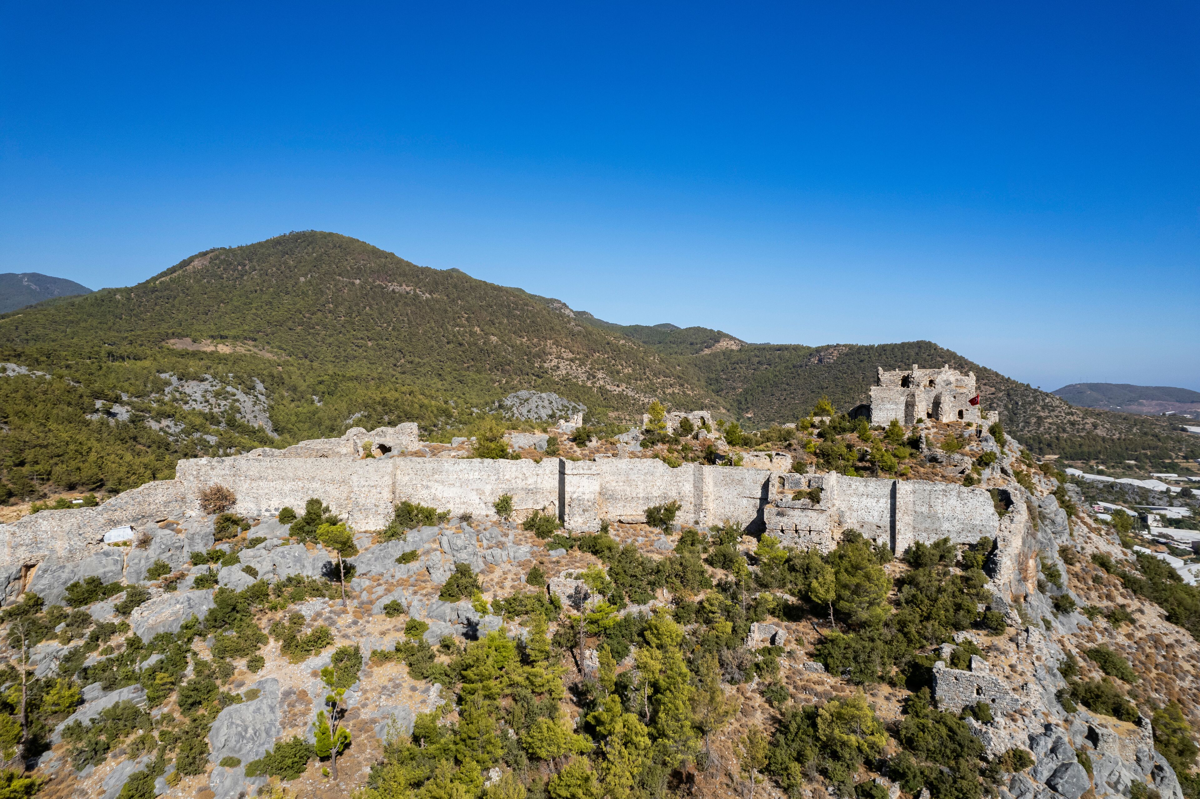 A historic stone fortress on a hill with surrounding greenery and mountains.