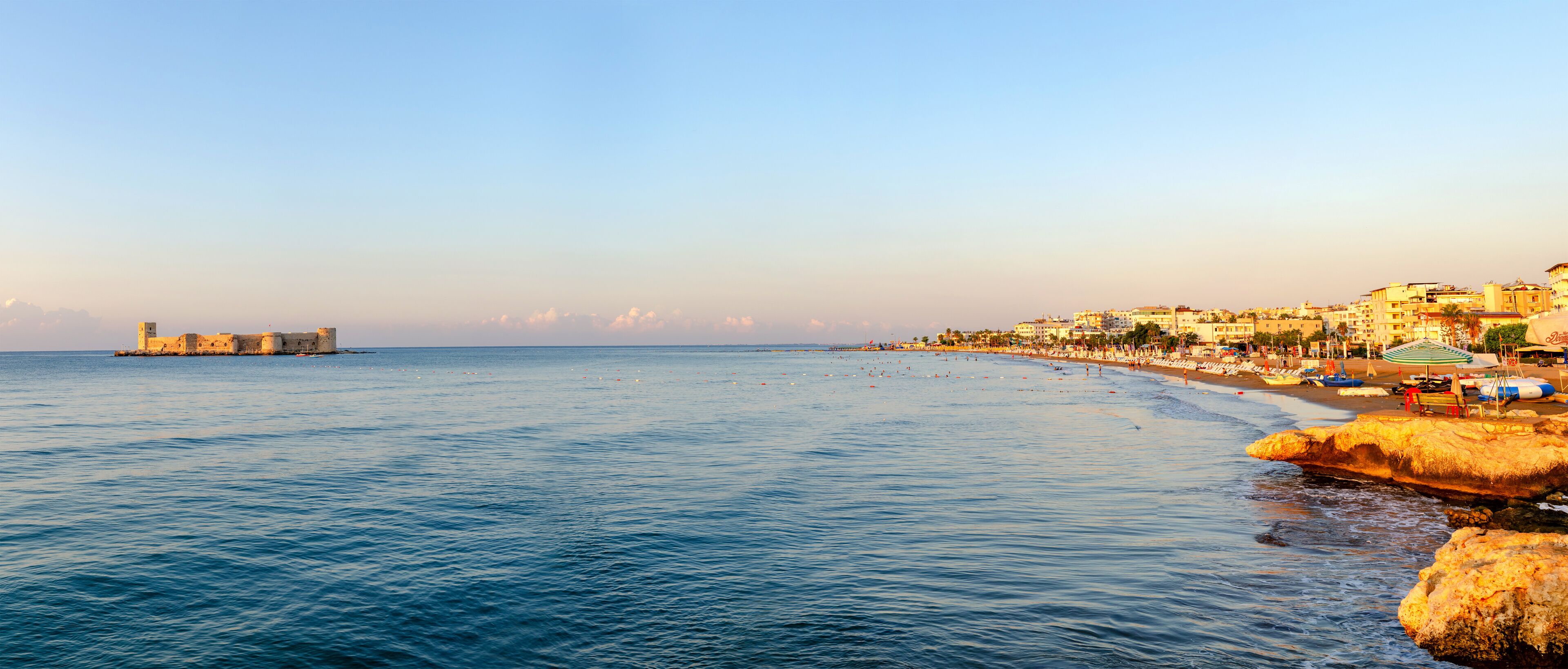A coastal town with a beach, rocky shoreline, and a distant fort.