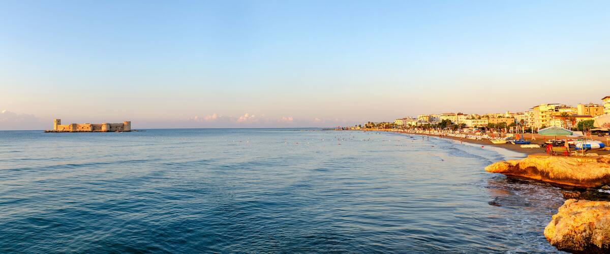 A coastal town with a beach, rocky shoreline, and a distant fort.