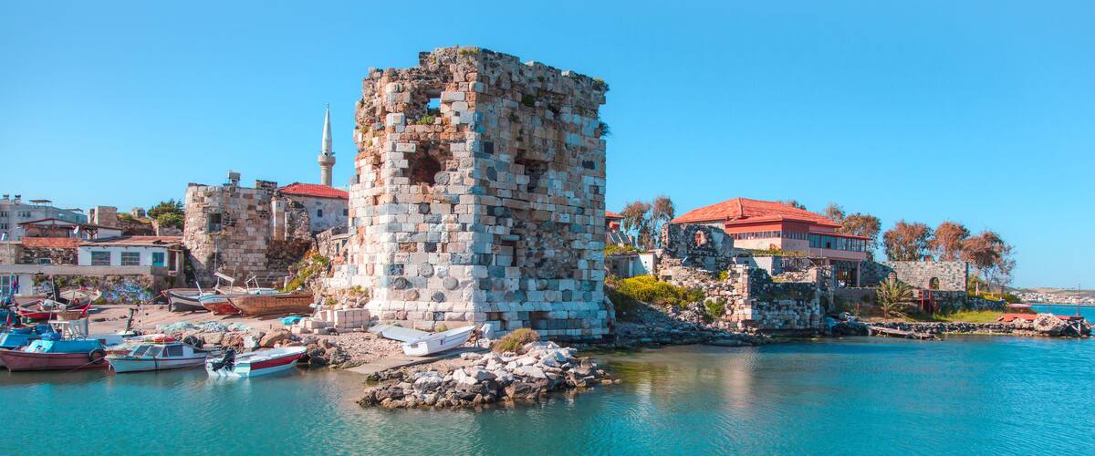 A historic stone tower by a waterfront with boats and buildings in the background.