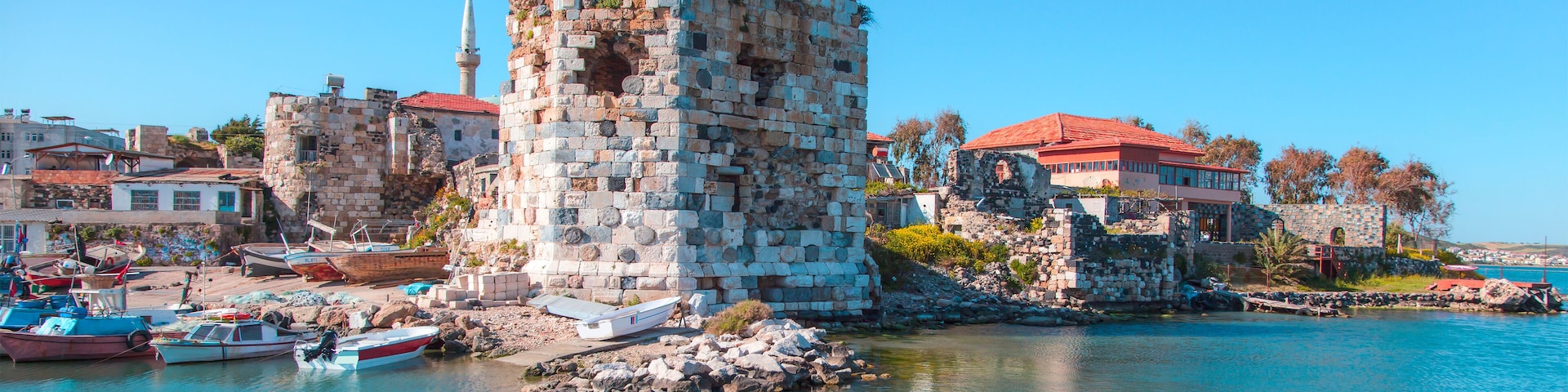 A historic stone tower by a waterfront with boats and buildings in the background.