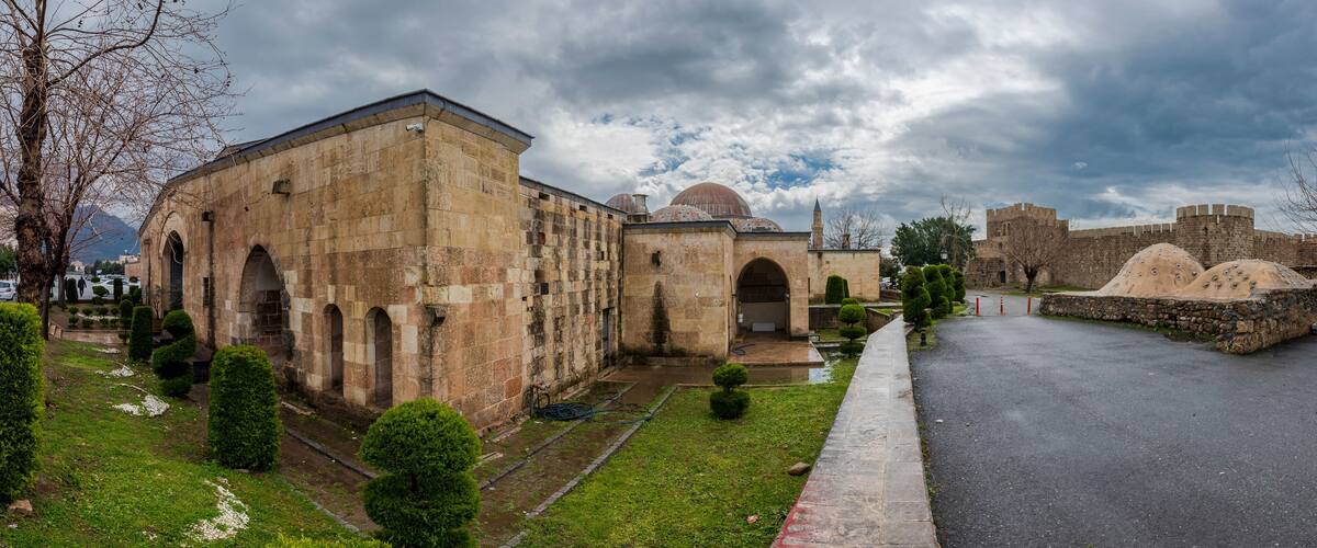 A historic stone building with arches and a dome, surrounded by well-maintained greenery and a paved road.