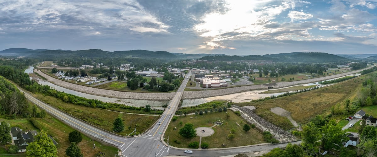 A river flowing through a town with a bridge, surrounded by hills and greenery.