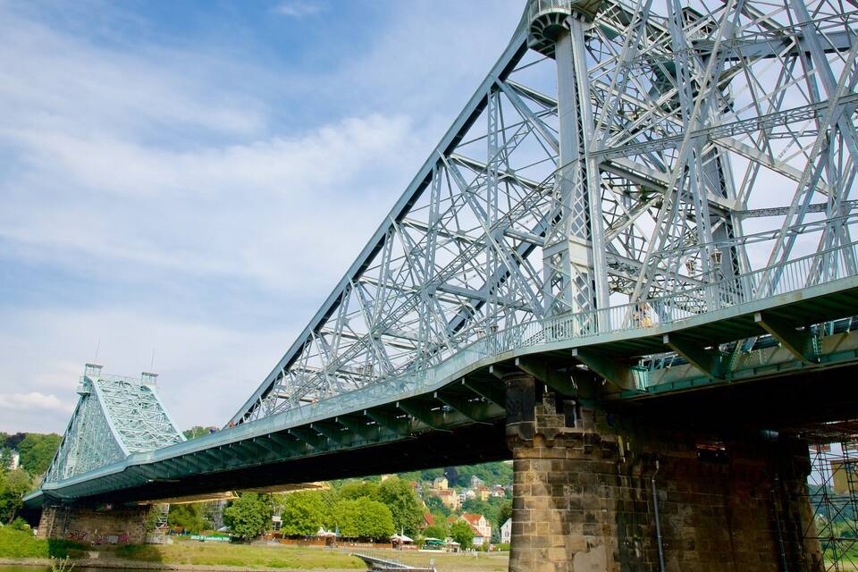 Un puente de acero con una pasarela verde, un río abajo y una vista distante de una torre y edificios.