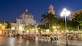A plaza with a large white building featuring domes and a bell tower, surrounded by palm trees and street lamps.