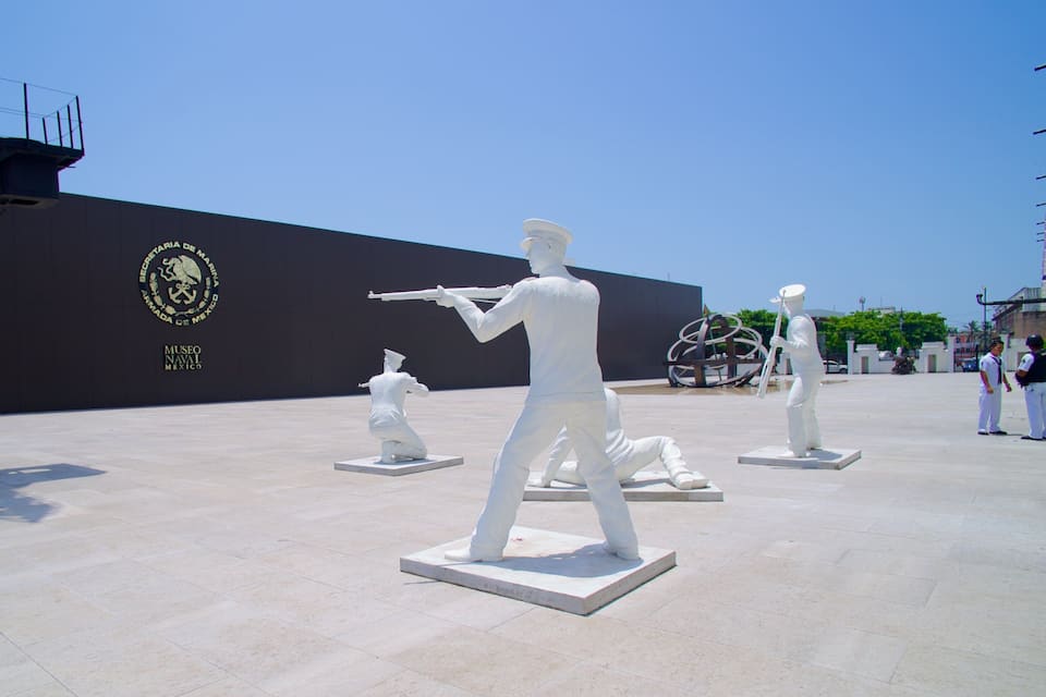 Un museo naval con estatuas blancas de marineros y un gran emblema en la pared.