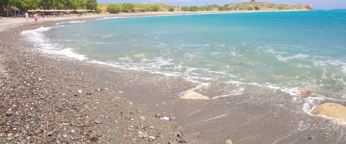 A pebbly beach with clear turquoise water, a distant hill, and a few people walking along the shore.