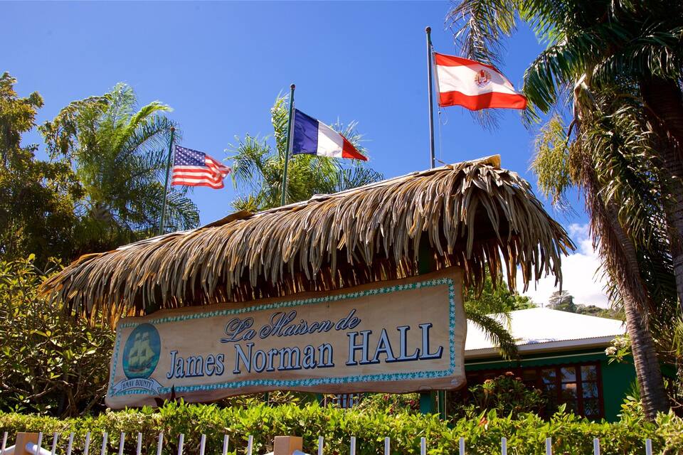 A thatched-roof building with flags, including the American and French flags, and a sign reading "James Norman Hall".