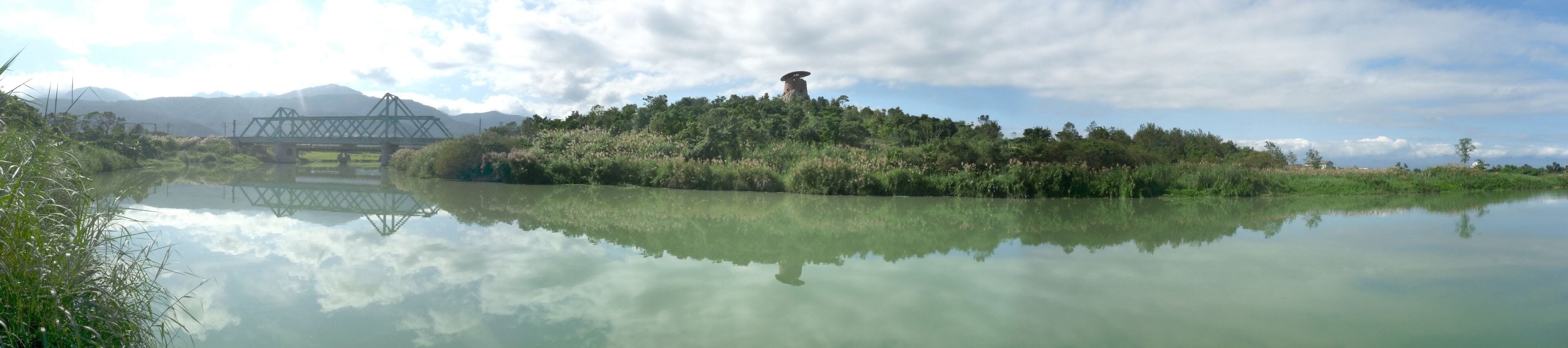 A calm lake reflecting a bridge and a hill with a structure on top.
