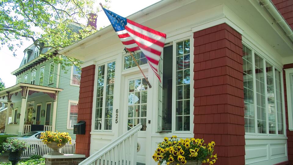 A house with a red brick exterior, white trim, and a front porch with a white railing. There are two flower pots with yellow flowers on the porch. An American flag is hanging on the front of the house.