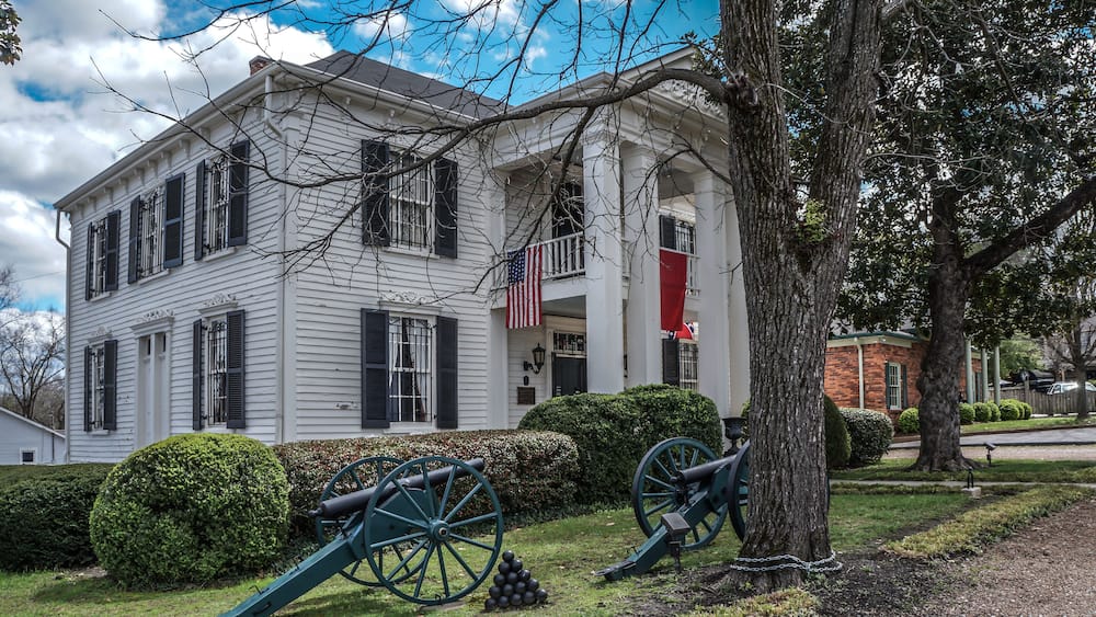 A white colonial house with black shutters, an American flag, and two cannons in the front yard.