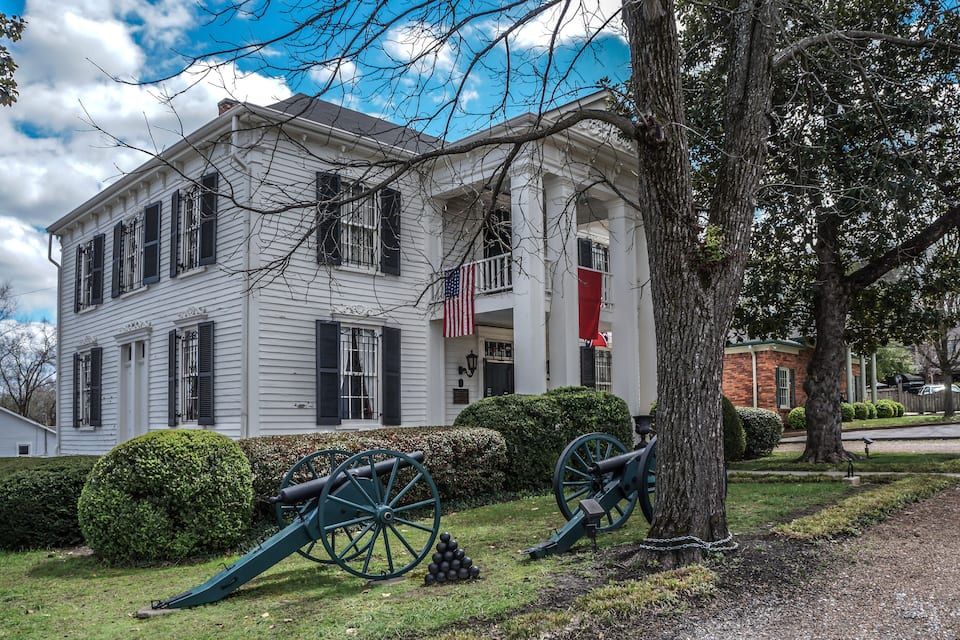 A white colonial house with black shutters, an American flag, and two cannons in the front yard.
