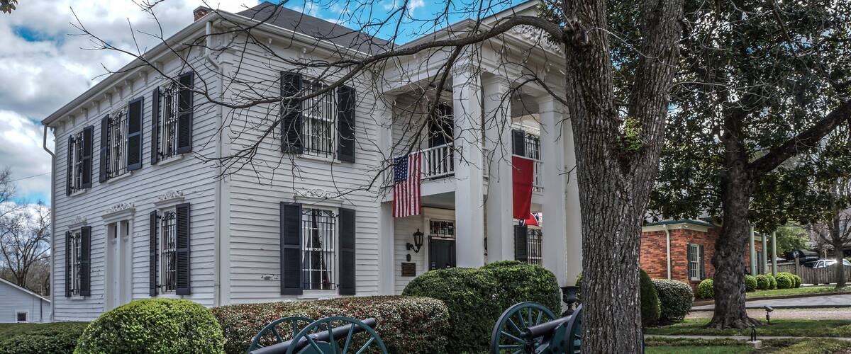 A white colonial house with black shutters, an American flag, and two cannons in the front yard.