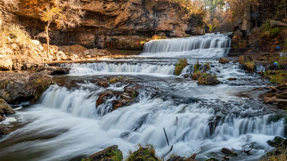 A multi-tiered waterfall surrounded by rocky terrain and autumn foliage.