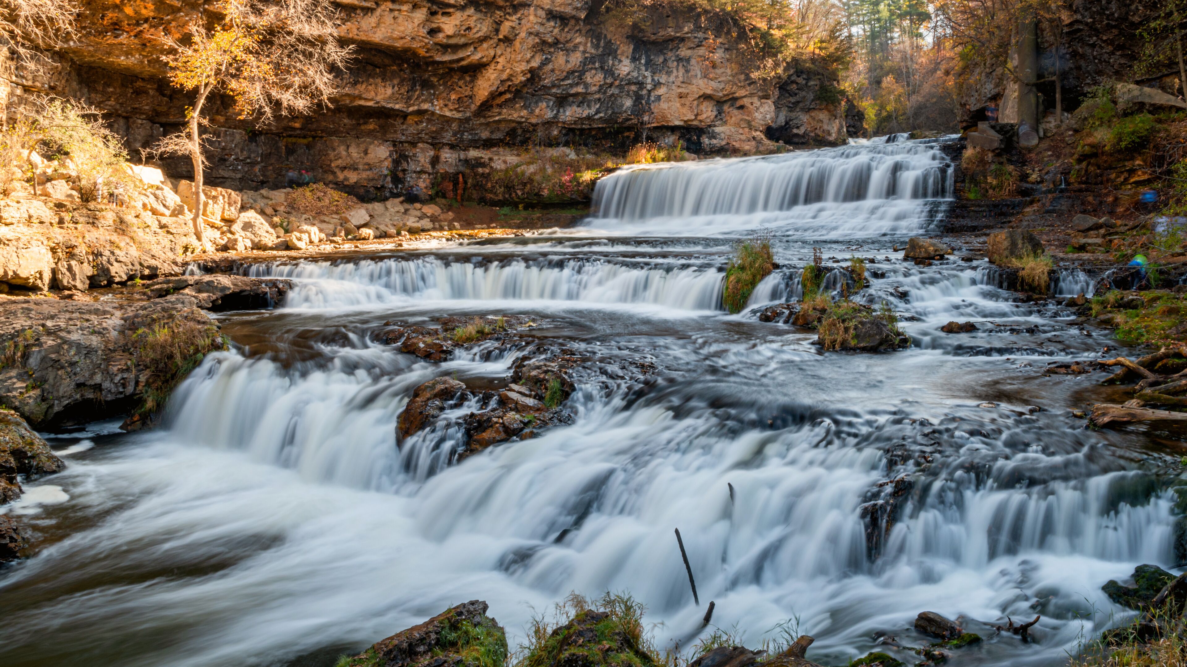 A multi-tiered waterfall surrounded by rocky terrain and autumn foliage.