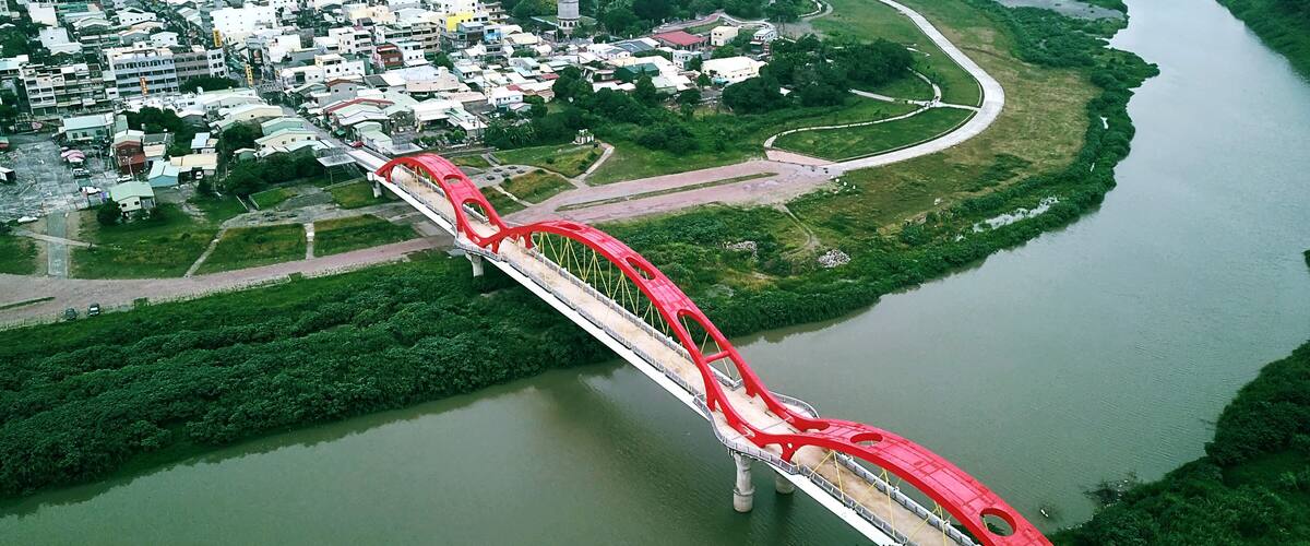 A red bridge spanning a river with a cityscape in the background.