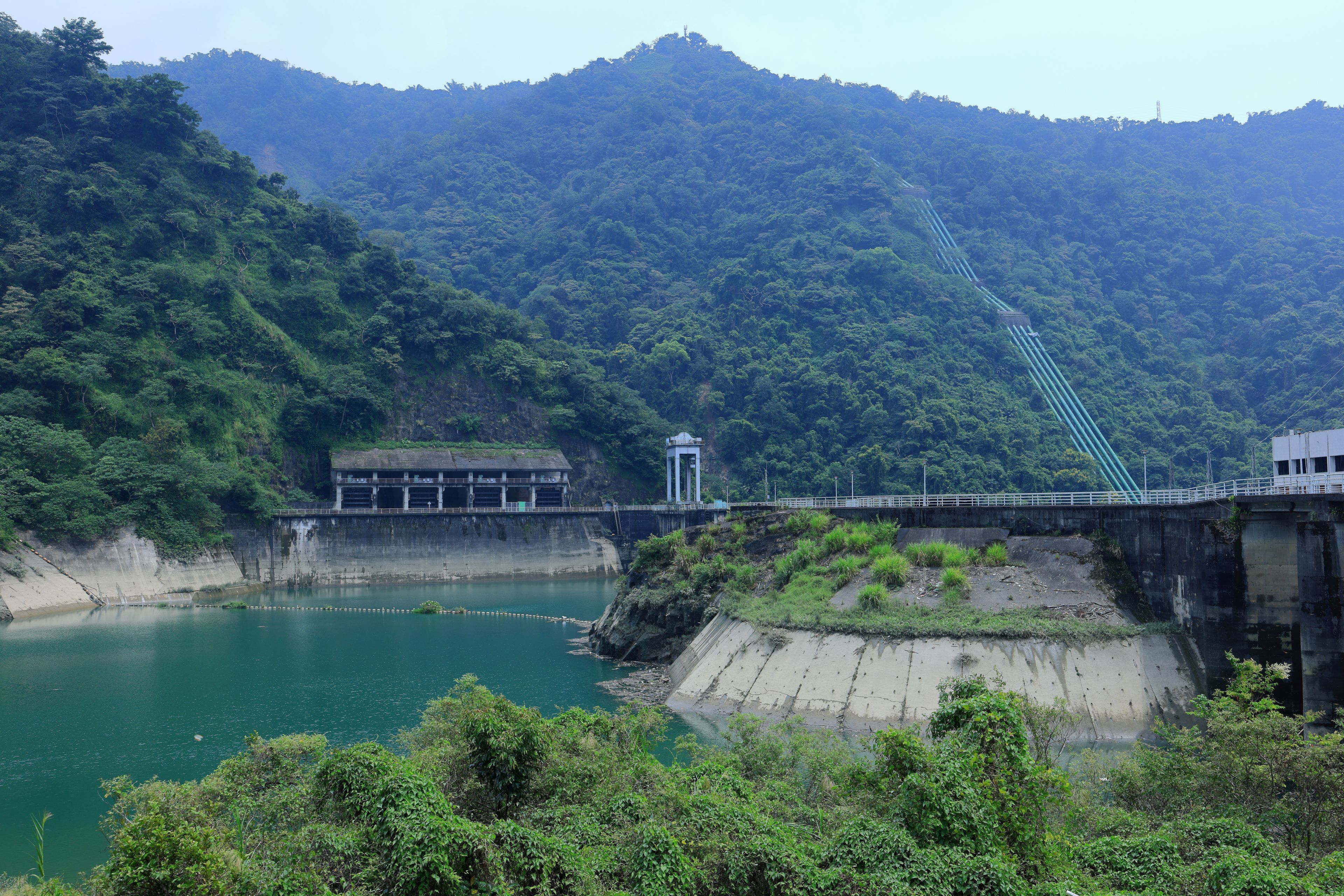 Uma barragem com uma ponte, um reservatório e montanhas ao fundo.