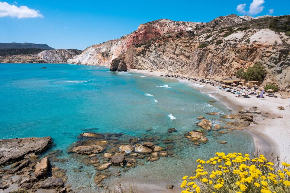 A coastal landscape with clear turquoise waters, rocky shorelines, and a sandy beach with umbrellas.