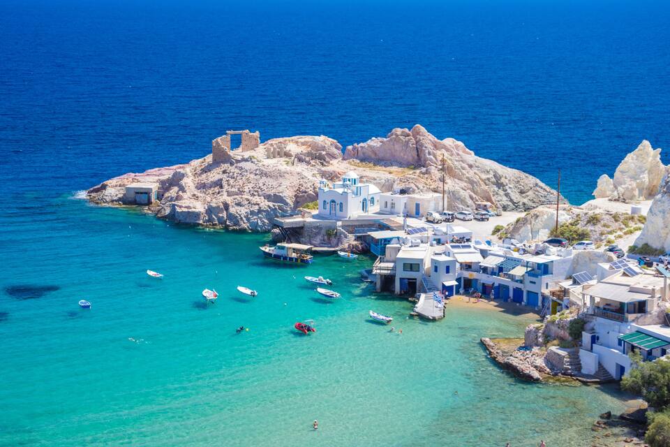A coastal village with white buildings, boats, and clear turquoise waters.