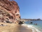 A beach with red cliffs, clear blue water, and a few people swimming.