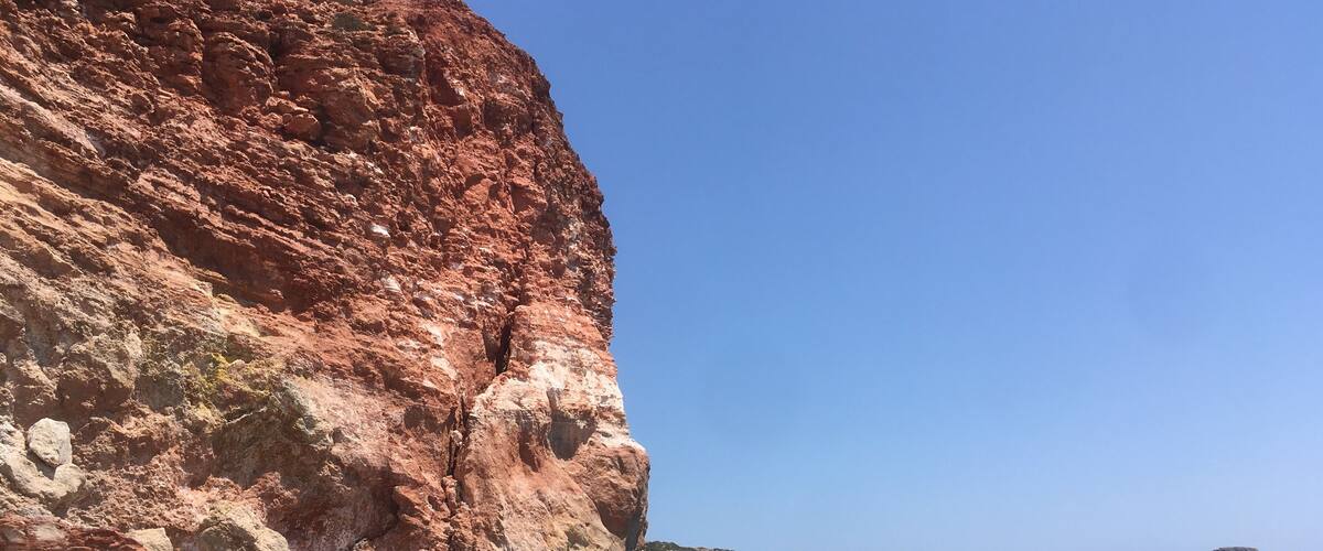 A beach with red cliffs, clear blue water, and a few people swimming.