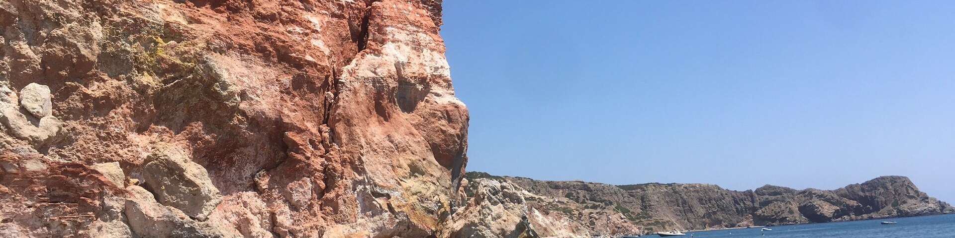 A beach with red cliffs, clear blue water, and a few people swimming.