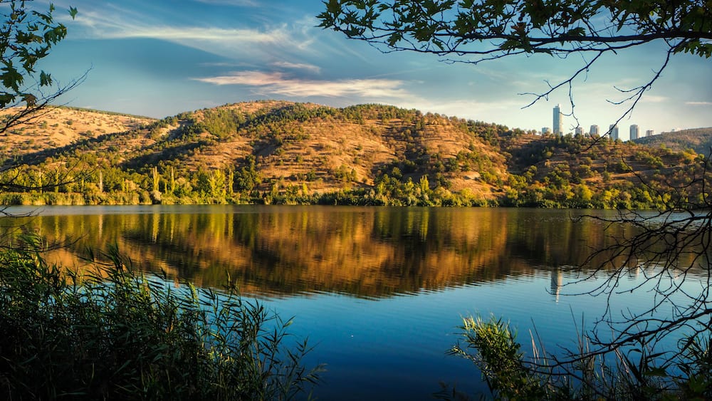 A calm lake reflecting the surrounding hills and a distant city skyline.