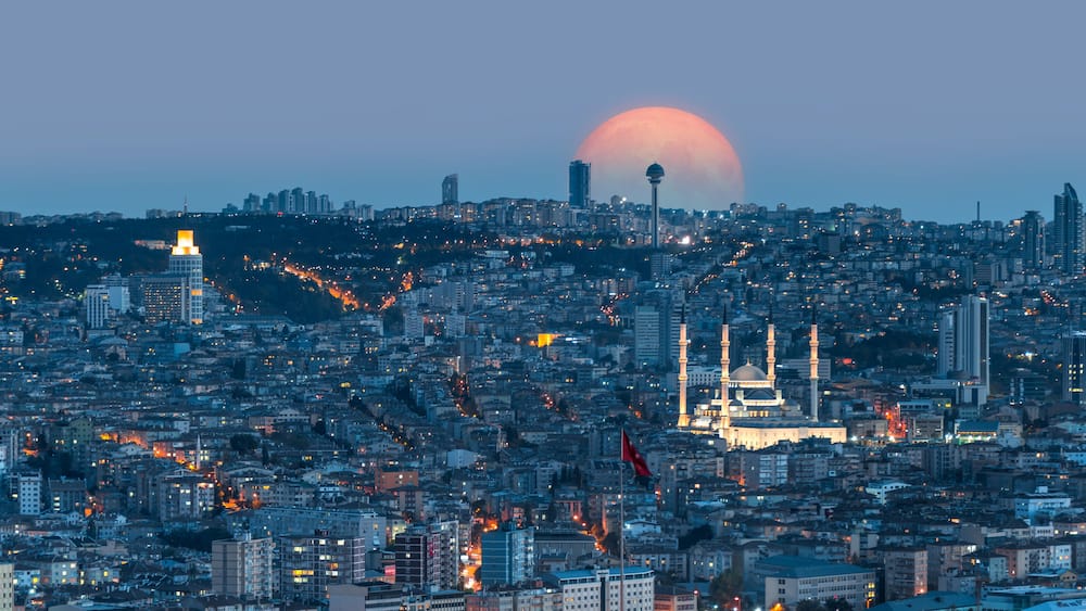 A cityscape with a prominent mosque and a large crescent moon in the sky.