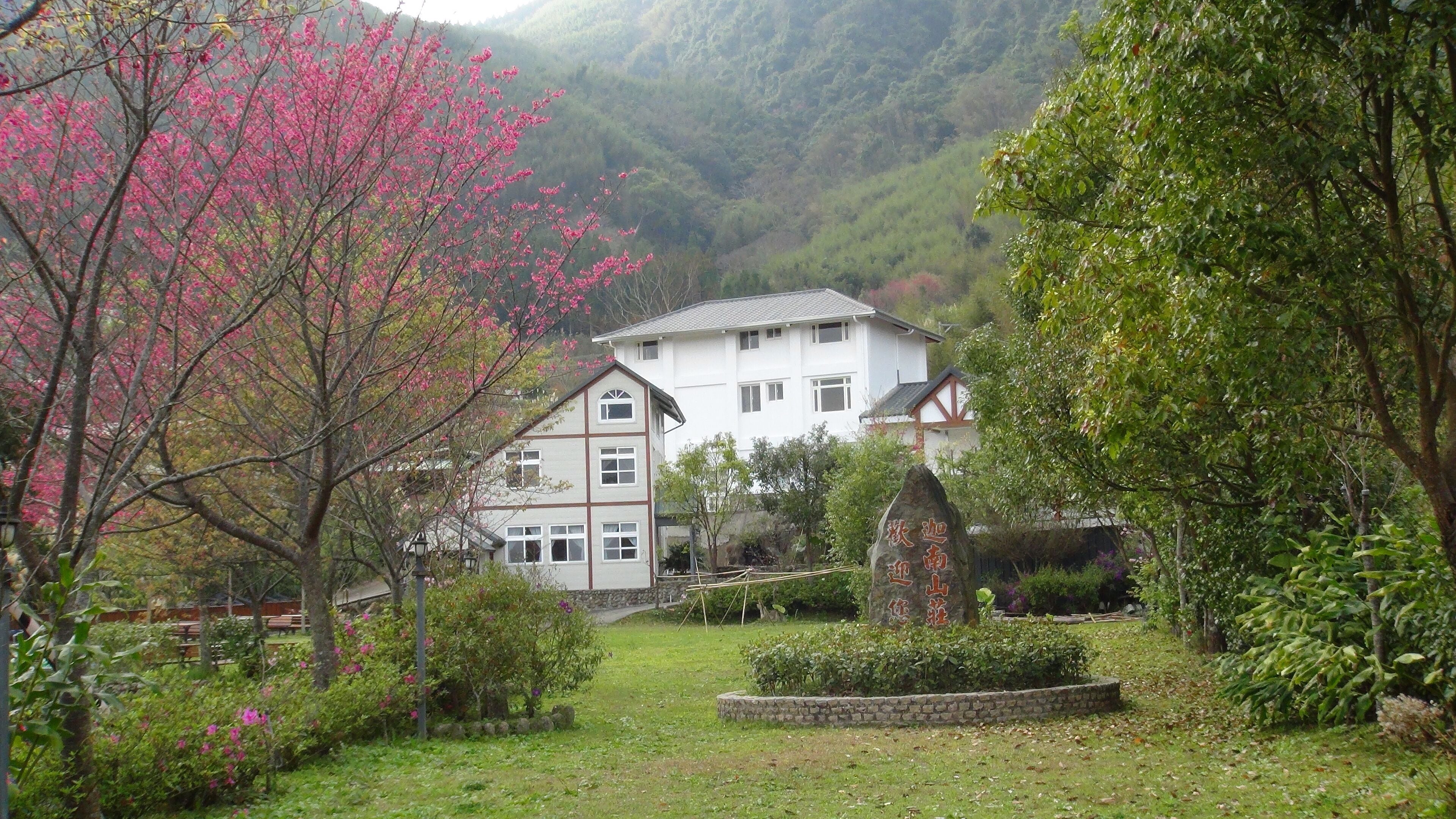 Una casa blanca rodeada de exuberante vegetación y árboles con flores rosadas en una zona montañosa.