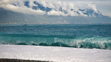 A coastal scene with waves crashing onto the shore, a distant mountain range partially obscured by clouds, and a few boats on the water.