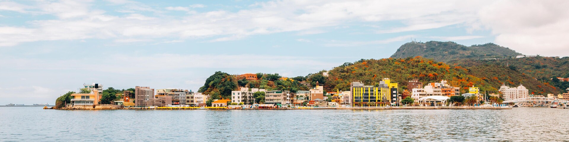 Uma cidade costeira com edifícios coloridos ao longo da orla, uma colina com folhagem outonal e um céu claro.