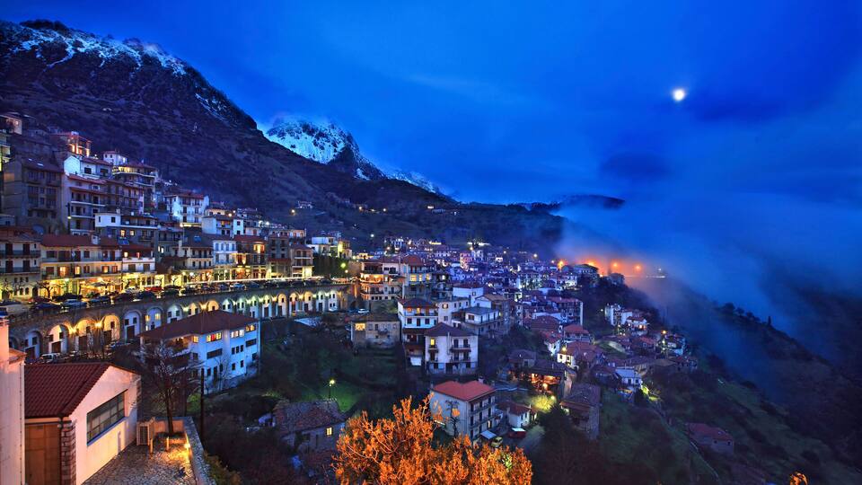 A mountainous town at dusk with snow-capped peaks and illuminated buildings.