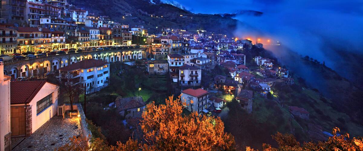 A mountainous town at dusk with snow-capped peaks and illuminated buildings.