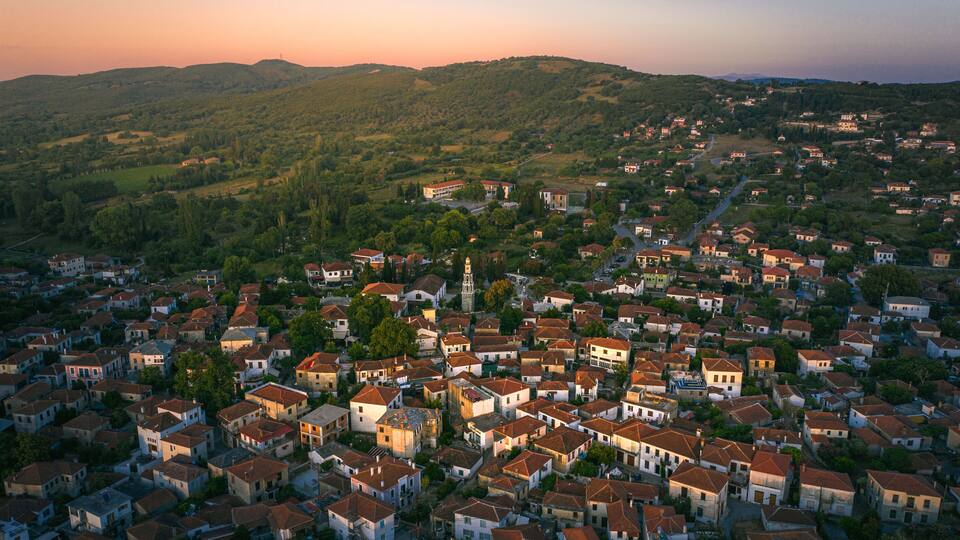 A densely packed town with red-roofed houses, surrounded by green hills and a clear sky.