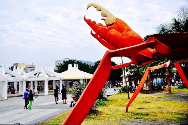 Eine große, rote Krabskulptur auf einem öffentlichen Platz, mit Passanten und einem weißen Gebäude im Hintergrund.